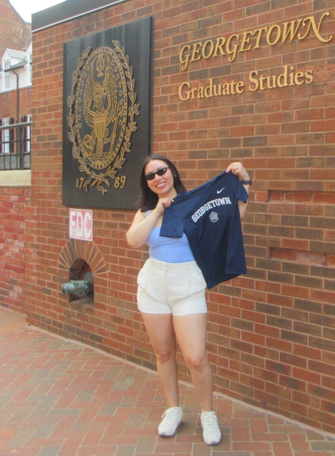 Salvadoran woman standing in front of the Georgetown University Graduate Studies sign. She is smiling and holding up a Georgetown University t-shirt.